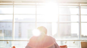 A man sitting in an airport facing away from the camera in front of a large window with sun streaming in towards the viewer
